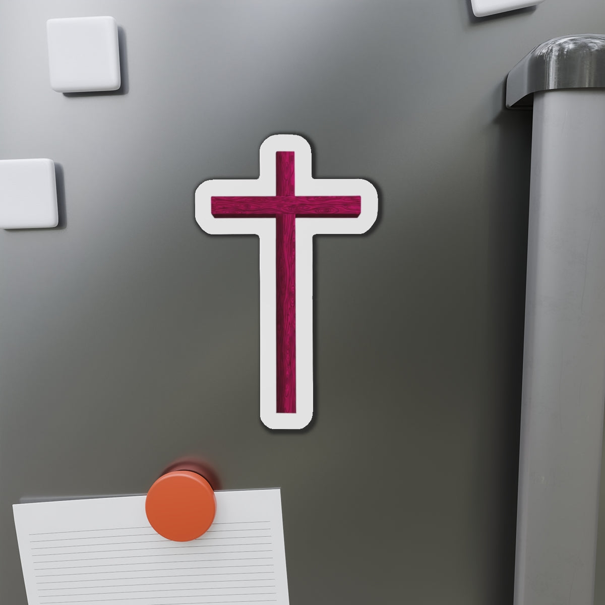 Cross-shaped magnet on a refrigerator door with other magnets and a pen holder.